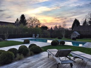 Coucher de soleil sur un espace piscine avec son habillage de terrasses bois et pierre bleue. La touche « cosy » étant apportée par les végétaux proches.